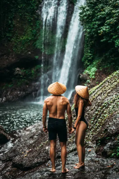 Photo of Couple admiring a beautiful waterfall in Indonesia. Tourists at the waterfall, rear view. Couple on vacation in Bali. Honeymoon trip. A couple in love travels the world. Vacation on the island of Bali