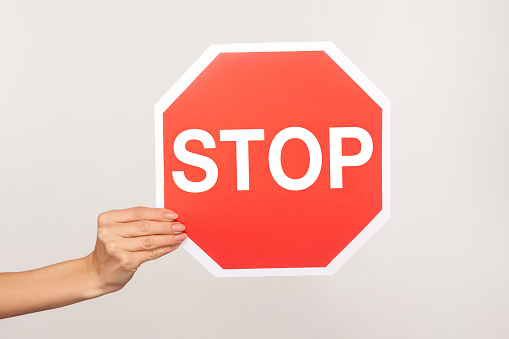 Closeup of female hand holding octagonal Stop symbol, ban concept, arm showing red traffic sign warning of forbidden way, restricted area, prohibited to go. studio shot isolated on blue background