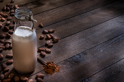 Front view of a bottle full of chocolate milkshake surronded by cocoa beans and a teaspoon with cocoa powder on a rustic wooden table. The objects are at the left side of the image leaving a useful copy space at the right side. Low key DSLR photo taken with Canon EOS 6D Mark II and Canon EF 24-105 mm f/4L