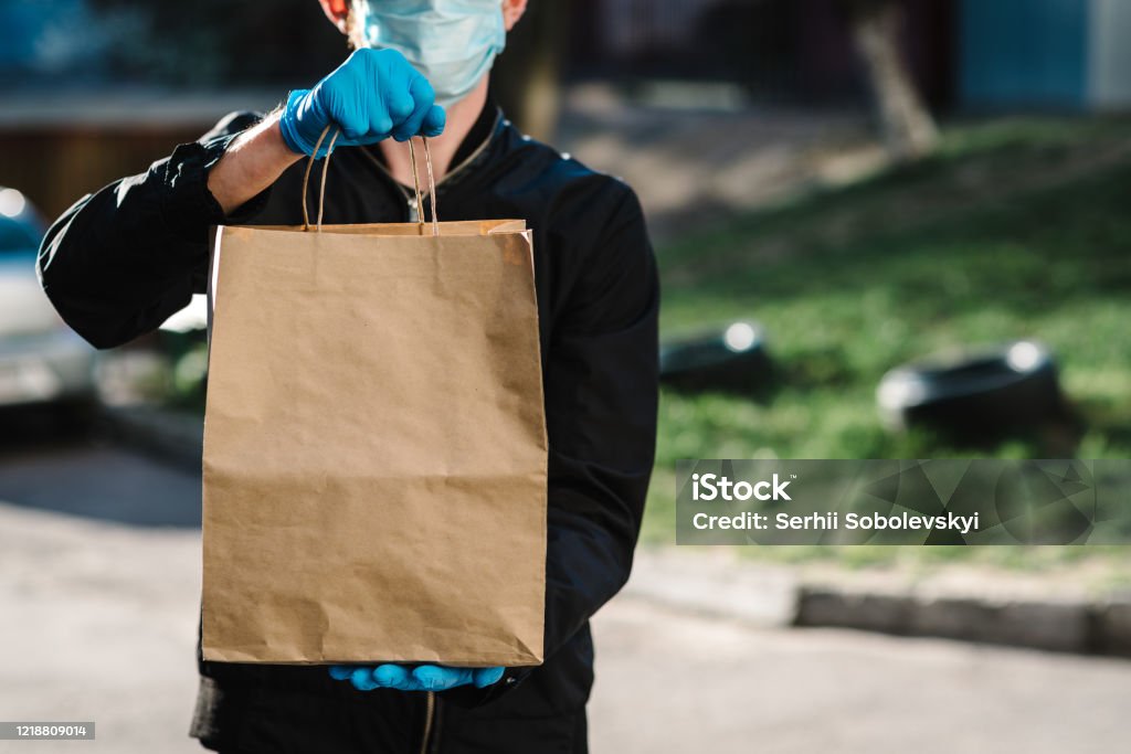 Courier in protective mask, medical gloves delivers takeaway food.  Employee hold cardboard package. Place for text. Delivery service under quarantine, 2019-ncov, pandemic coronavirus, covid-19. Delivering Stock Photo