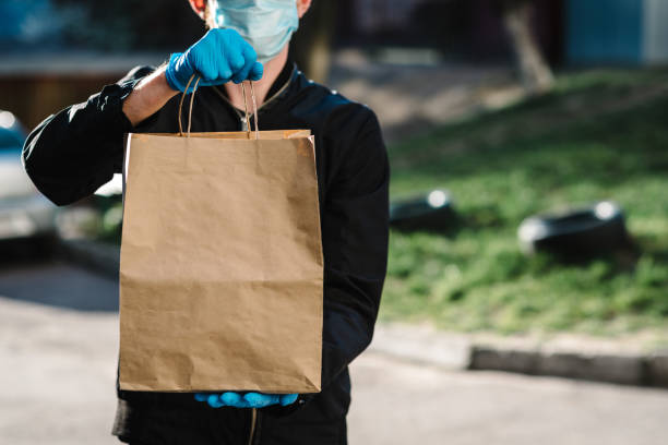 Courier in protective mask, medical gloves delivers takeaway food.  Employee hold cardboard package. Place for text. Delivery service under quarantine, 2019-ncov, pandemic coronavirus, covid-19. Courier in protective mask, medical gloves delivers takeaway food.  Employee hold cardboard package. Place for text. Delivery service under quarantine, 2019-ncov, pandemic coronavirus, covid-19. take out food photos stock pictures, royalty-free photos & images