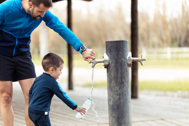 drôle petit garçon remplissant sa bouteille d’eau dans la fontaine publique tandis que son père l’aide. - water child bottle little boys photos et images de collection