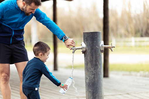Shot of funny little boy filling his water bottle in public fountain while his father helps him.