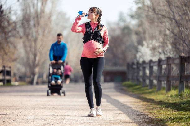 pregnant young woman drinking water in the park. in the background, her husband walking his son with the cart. - relaxation exercise child mother human pregnancy imagens e fotografias de stock