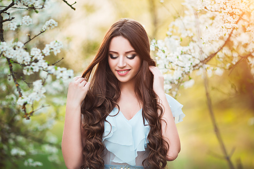 Happy beautiful young woman with long black healthy hair enjoy fresh flowers and sun light in blossom park at sunset