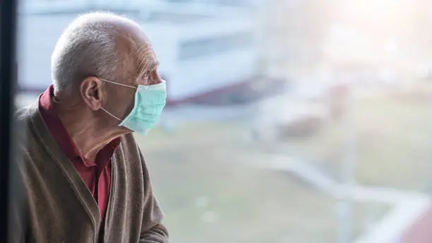 aged pensioner man with gray hair wearing medical facemask looking through window health care concept