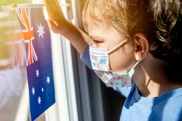 オーストラリアの旗を持つ外科用マスクを使って窓越しに見ている小さな子供の男の子 - sadness child little boys loneliness ストックフォトと画像