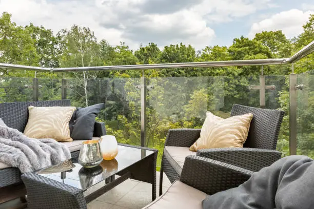 Stylish balcony with elegant rattan furniture and glass walls and green forest view