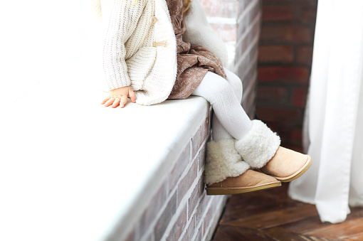 Faceless little girl in warm clothes and footwear sitting on the windowsill