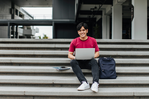 Portrait of young Asian man student sitting on stairs and using laptop.