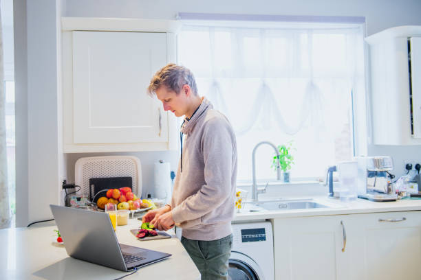 istock Young caucasian man working from home. Freelancer cooking vegetarian lunch on the kitchen table and working on pc laptop. Home office, remote workplace concept. Soft selective focus. Copy space. 1218841672