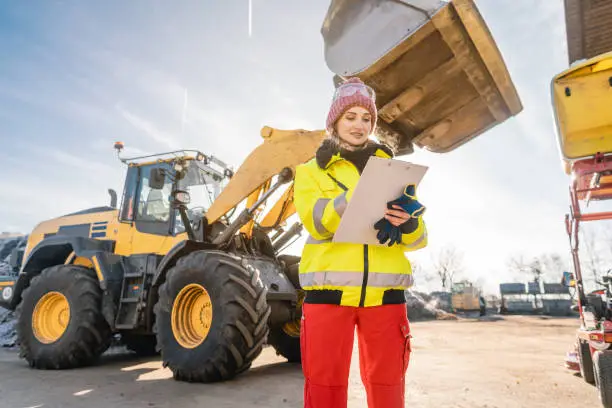 Woman with clipboard in front of construction machinery writing