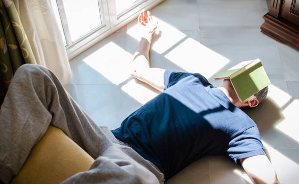 Young man resting on the floor whit a book on his face while enjoying the sun coming through the window. Concept of stay at home, freedom, boredom... Young man resting on the floor whit a book on his face while enjoying the sun coming through the window. Concept of stay at home, freedom, boredom... couch potato photos stock pictures, royalty-free photos & images