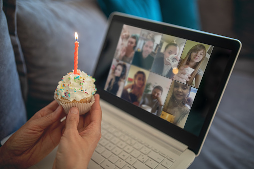 Mature woman celebrating her birthday alone at home but with friends online on video conference. She is sitting cozy on sofa in living room and holding cupcake with candle. She is lonely in quarantine during lockdown because of corona virus outbreak