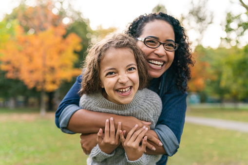 A mixed-race black mother is spending time with her elementary age daughter. The two individuals are bonding while enjoying time outdoors. The mother is standing behind her daughter and has her wrapped in a big hug. Both individuals are laughing. The child is looking directly at the camera. It is Autumn and trees with orange leaves are visible in the background.