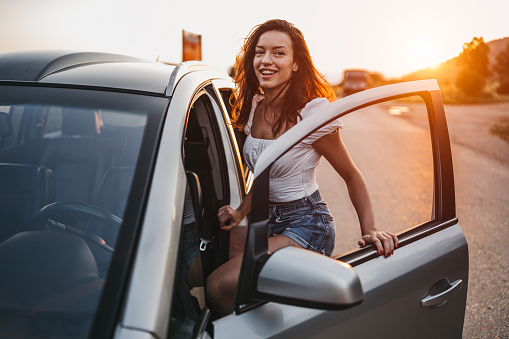 Beautiful young woman getting into car on country road at sunset