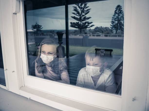 COVID-19 Outbreak. Sad sick children with face mask looking through the window during quarantine at home. Coronavirus isolation and kids mental health.