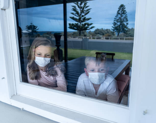 istock COVID-19 Outbreak. Sad sick children with face mask looking through the window during quarantine at home. Coronavirus isolation and kids mental health. 1218899707