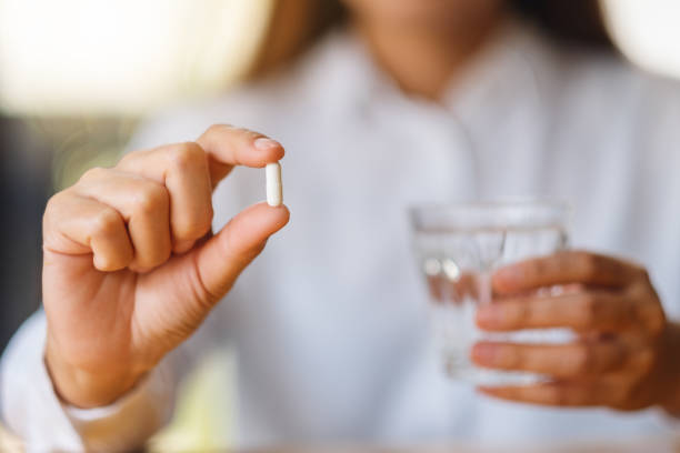 uma mulher segurando uma pílula branca e um copo de água - capsule pill white nutritional supplement - fotografias e filmes do acervo