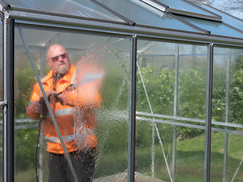 Construction worker cleaning filth with high pressure cleaner from a glass greenhouse.