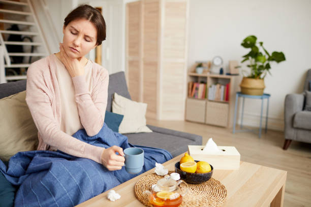 Woman has a sore throat Sick woman sitting on sofa she has a sore throat and she drinking tea with lemon to relieve the symptoms at home sore throat stock pictures, royalty-free photos & images