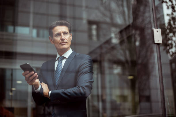 German businessman Formally dressed 55-year-old businessman is standing in the bright office and using smartphone. georgijevic frankfurt stock pictures, royalty-free photos & images