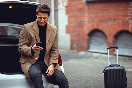 55-year old handsome man is sitting on the edge of the car trunk and using smartphone. There is a suitcase next to him.