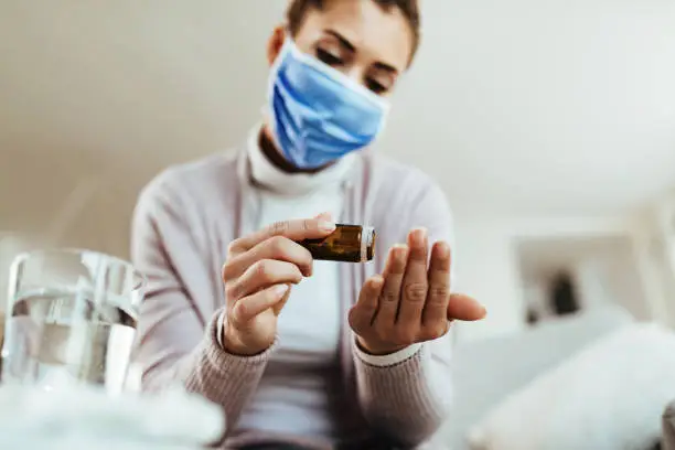 Photo of Close-up of woman with face mask taking antiviral drugs at home.