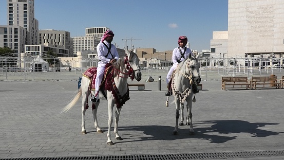 Doha, Qatar - February 20, 2019: two heritage Police Officers in traditional 1940s Qatari uniform riding white Arabian Horses at square of Souq Waqif. Popular tourist attraction in Doha city center.