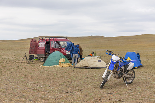Tourists camping in Mongolian hills. Three tents under the open cloudy sky. A motorcycle in the foreground.
