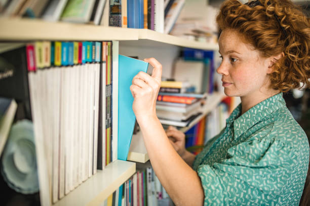 istock Redhead woman taking a book from the shelf in a library. 1218927151