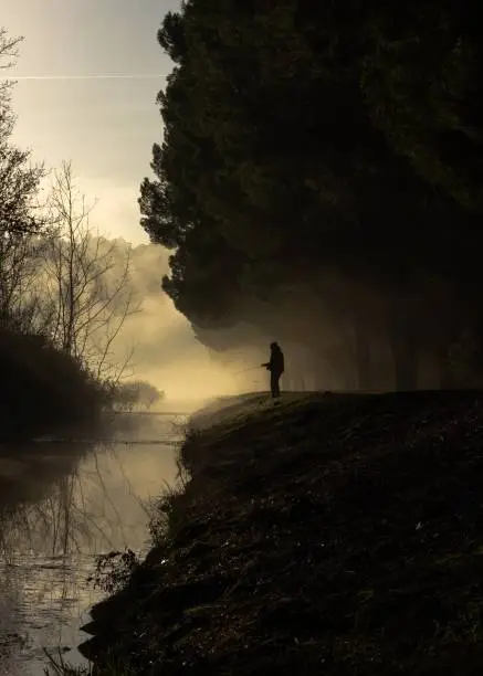 Person fishing in a small lake near a forest with fog at the sunrise