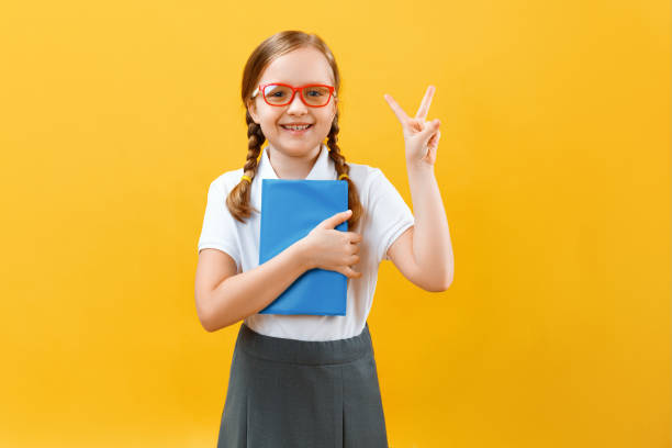 黄色の背景に眼鏡をかけた小さな女子高生の肖像画。子供は手に本を持ち、勝利のサインを見せる。教育、成功、学校のコンセプト。スペースをコピーする ストックフォト