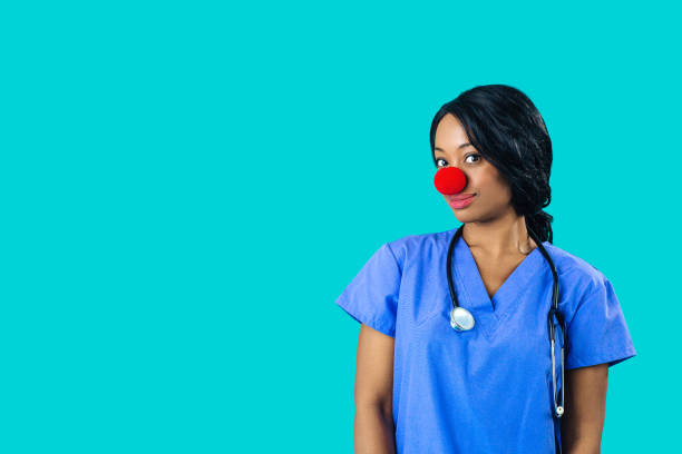 portrait of a smiling female doctor or nurse wearing blue scrubs uniform and red nose looking at camera isolated on blue background - clowns nose imagens e fotografias de stock