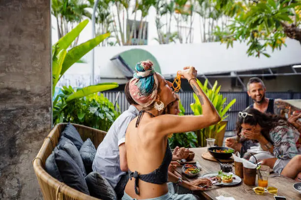 A wide shot of male and female friends eating a meal together in a food and drink establishment in Bali, Indonesia. The main focus is a woman feeding herself spaghetti using her hand.