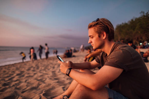 Relaxed Weekends on the Beach A side view shot of a young caucasian man sitting down on the beach, he is using a smart phone in Bali, Indonesia. kuta beach stock pictures, royalty-free photos & images