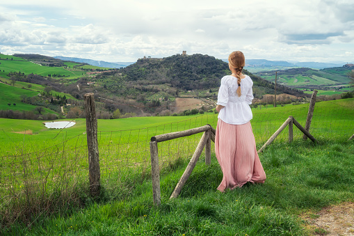 Rear view of a woman with vintage clothes admires the beauty of green rolling hills in the heart of Tuscany, Italy.