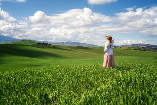 Rear view of a woman with vintage clothes admires the beauty of green rolling hills in the heart of Tuscany, Italy.