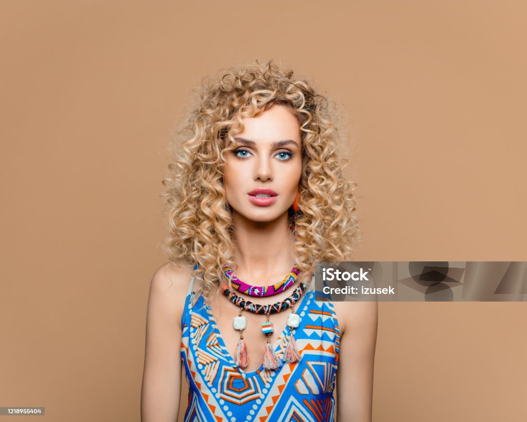 Boho girl in tribal fashion dress and jewelry against brown background Summer portrait of beautiful long curly blond hair young woman wearing boho style blue dress and tribal jewelry, smiling at camera. Studio shot on brown background. 25-29 Years Stock Photo