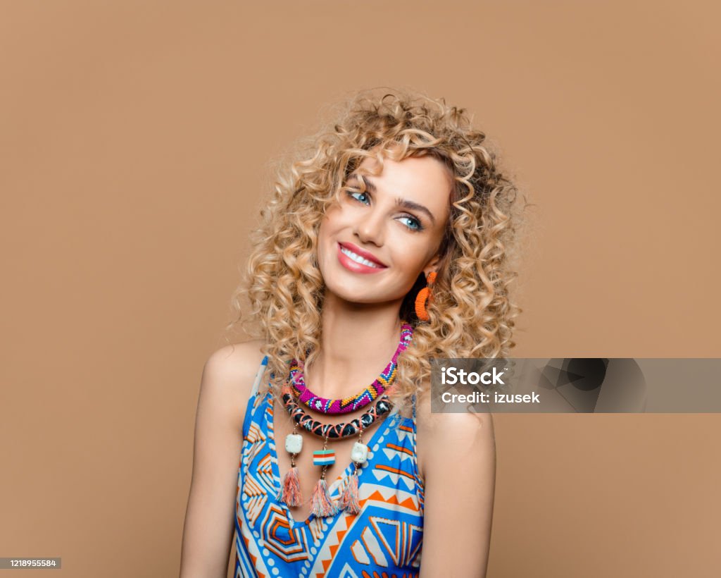 Cute woman in boho style outfit against brown background Summer portrait of beautiful long curly blond hair young woman wearing boho style dress and jewelry, looking away and smiling. Studio shot on brown background. Boho Stock Photo