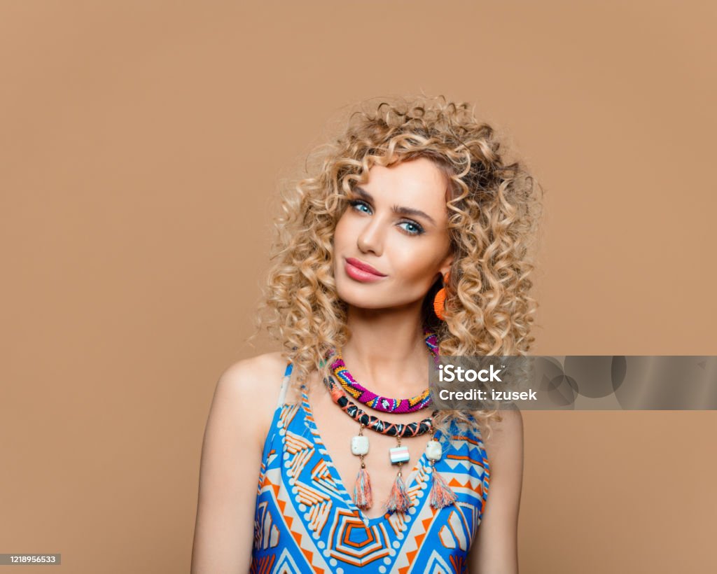 Fashion portrait of woman in boho style outfit Summer portrait of beautiful long curly hair young woman wearing boho style dress and jewelry, smiling at camera. Studio shot on brown background. Adult Stock Photo
