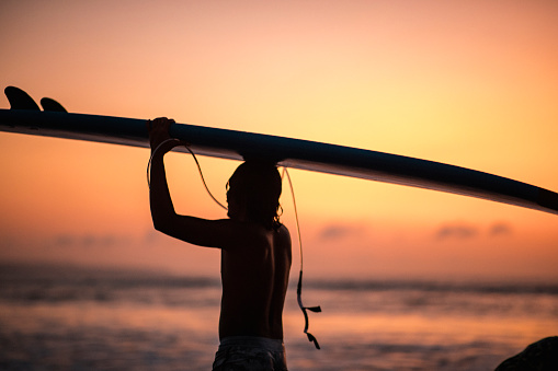 A silhouette of a man carrying a surfboard on his head and supporting it with his hands, he is getting ready to surf. He is at Kuta beach in Bali, Indonesia at dusk.