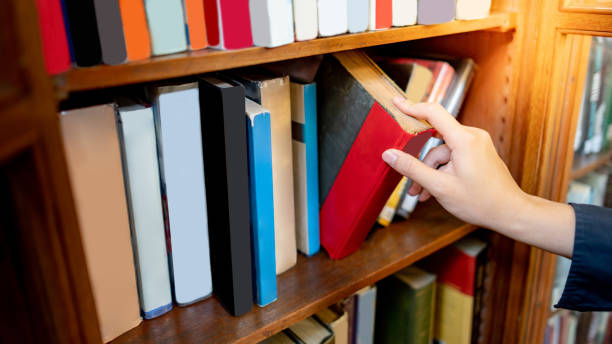 Male hand picking old book from bookshelf Male university student hand choosing and picking old book from old wooden bookshelf in college library. Antique textbook resources for education research. History, Law and Literature learning opportunity dictionary definition stock pictures, royalty-free photos & images