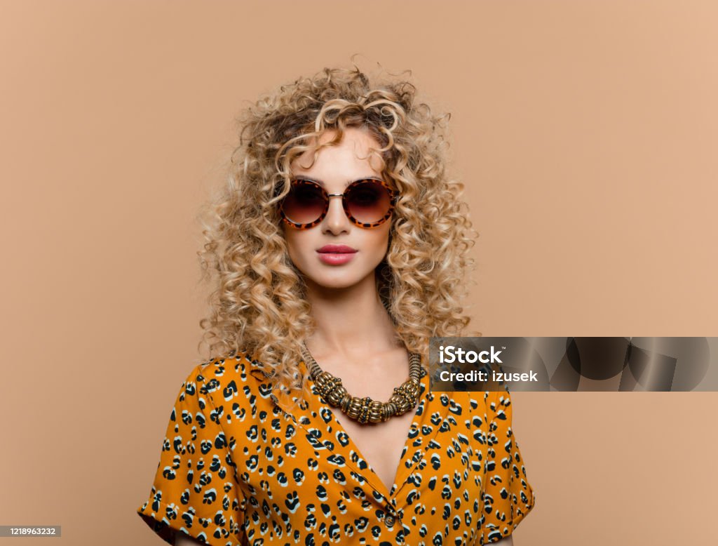 Fashion portrait of curly hair woman in leopard print dress Summer portrait of beautiful long curly hair young woman wearing leopard print dress, gold necklace and sunglasses, looking at camera. Studio shot on brown background. Glamour Stock Photo