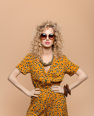 Summer portrait of beautiful long curly hair young woman wearing leopard print dress, gold necklace and sunglasses, looking at camera. Studio shot on brown background.