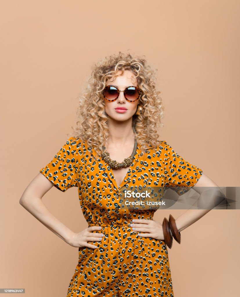 Fashion portrait of woman in leopard print dress Summer portrait of beautiful long curly hair young woman wearing leopard print dress, gold necklace and sunglasses, looking at camera. Studio shot on brown background. Women Stock Photo