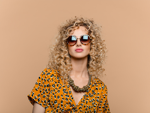 Fashion portrait of beautiful long curly hair young woman wearing leopard print dress, gold necklace and sunglasses, looking at camera. Studio shot on brown background.