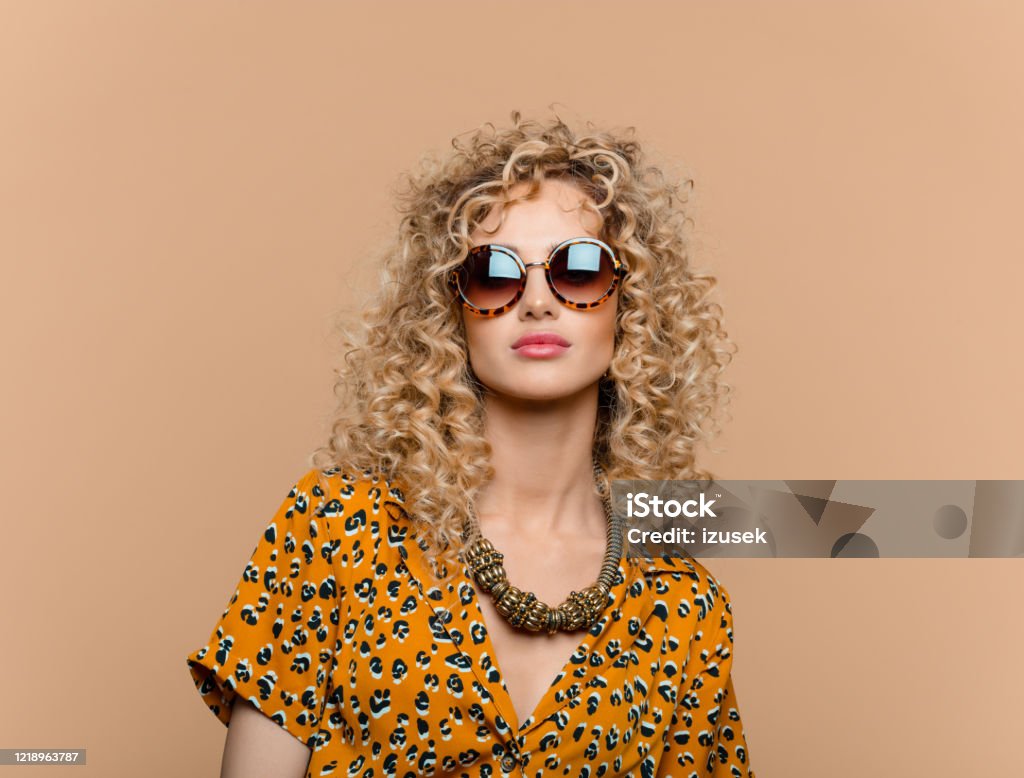 Summer portrait of curly hair woman in leopard print dress Fashion portrait of beautiful long curly hair young woman wearing leopard print dress, gold necklace and sunglasses, looking at camera. Studio shot on brown background. Sunglasses Stock Photo