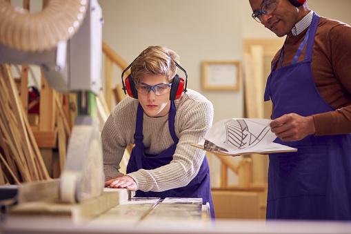 Tutor con estudiante de carpintería masculina en el taller de estudio para el aprendizaje en la universidad usando sierra de banco photo