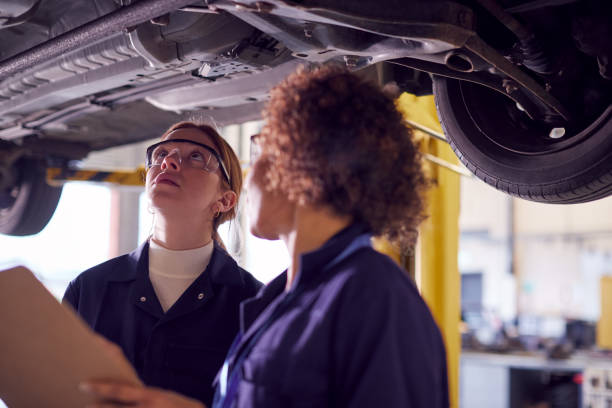 istock Female Tutor With Student Looking Underneath Car On Hydraulic Ramp On Auto Mechanic Course 1218971805